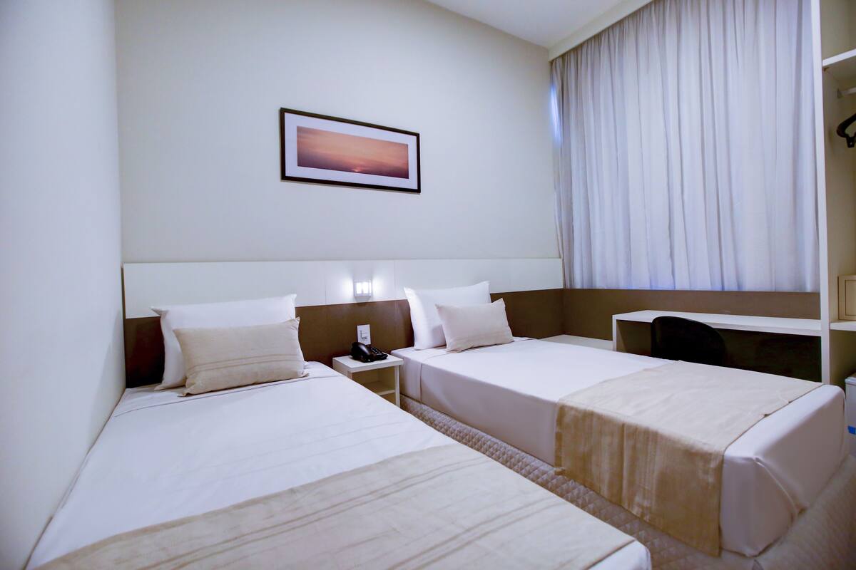 A hotel room with two beds, a bedside table with a phone, a nightstand, a framed picture on the wall, and a large window with curtains.
