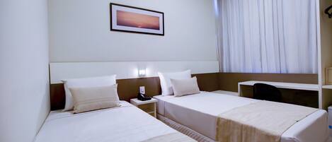 Habitación de hotel con dos camas, una mesita de noche con teléfono, una mesita auxiliar, un cuadro en la pared y un ventanal con cortinas.