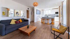 Un salon moderne avec un canapé, une table basse et un fauteuil. On aperçoit une cuisine à l’arrière-plan, avec un comptoir-bar et des tabourets.