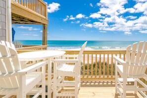 Een houten terras met witte stoelen en een tafel, uitkijkend op een strand.