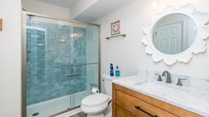 Een badkamer met een douchecabine van glas, een wit toilet, een houten wastafelmeubel met een marmeren werkblad, en een ronde spiegel met een getande rand.