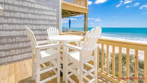 Een houten terras met een witte tafel en stoelen, uitkijkend op een strand met helderblauw water en een houten balustrade.