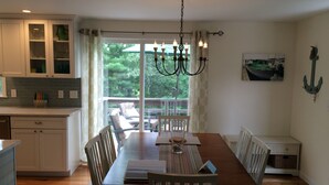 A dining area with a wooden table, chairs, and a chandelier. There is a window with curtains, a view of greenery outside, and a painting of a boat on the wall. - Updated Vacation Home Close to Town and Beaches (2221) (Orleans)