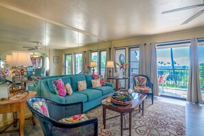 A spacious living room with a teal sofa, a dining area, and a balcony with a view.