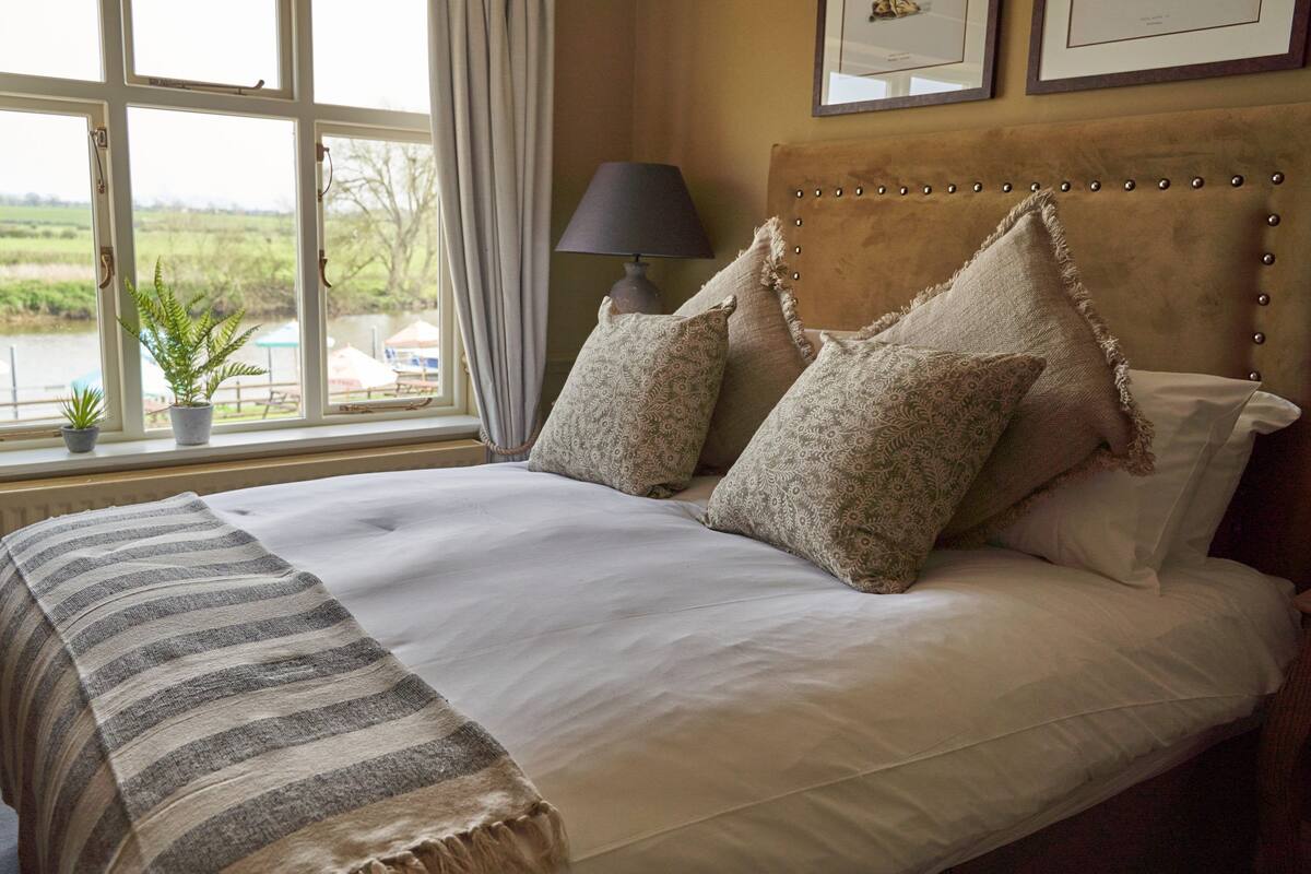 A bed with patterned pillows, a striped throw, and a view of a body of water through a window.