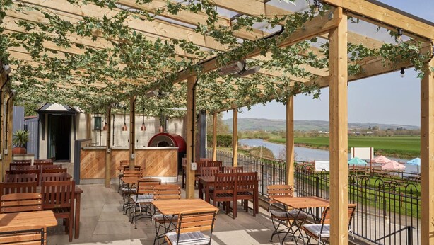 An outdoor patio with wooden tables and chairs, a barbecue area, and a view of a river and fields. - The Fleet (Tewkesbury)