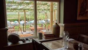 A dining area with a table set for two, a view of the outdoors through large windows, and a rustic decor including a large vase and a pitcher. - The Fleet (Tewkesbury)