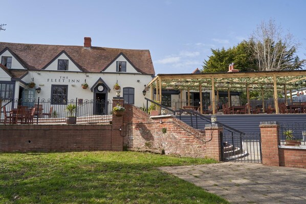 A traditional building with a sign reading "The Fleet Inn," featuring a brick facade, a covered outdoor seating area, and a staircase leading to the entrance. - The Fleet (Tewkesbury)
