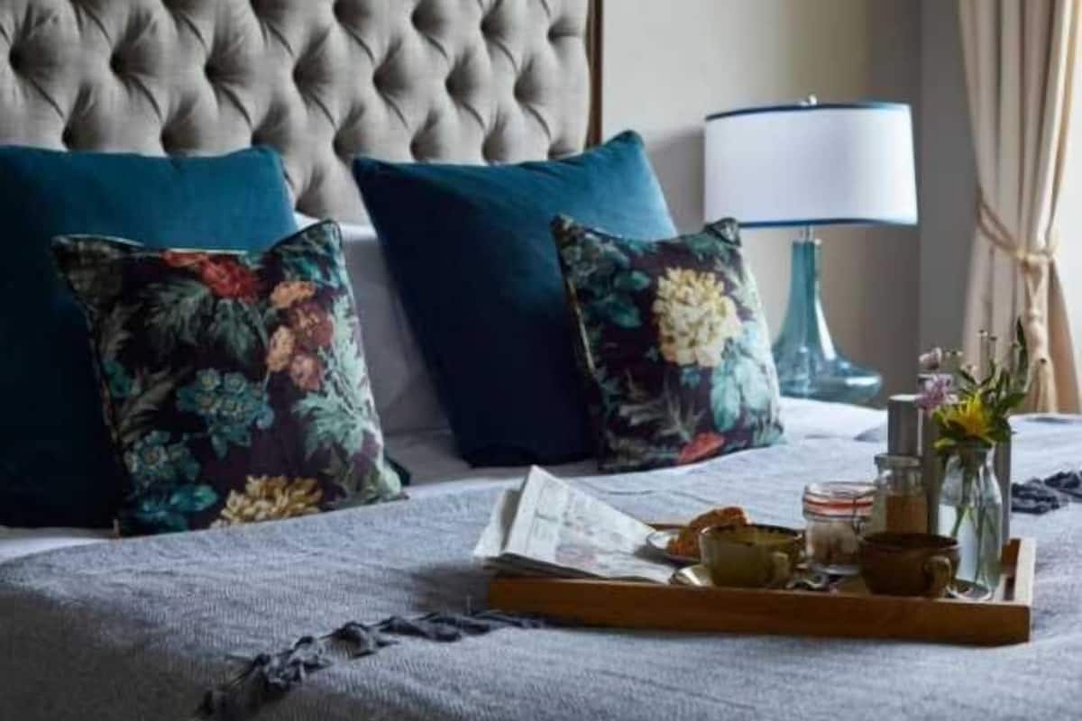 A bed with a tufted headboard, two floral-patterned pillows, a bedside table with a lamp, and a tray with a book, a cup, and a vase with flowers.