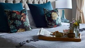 A neatly made bed with floral pillows, a bedside table with a lamp, and a tray with breakfast items. - The Fleet (Tewkesbury)
