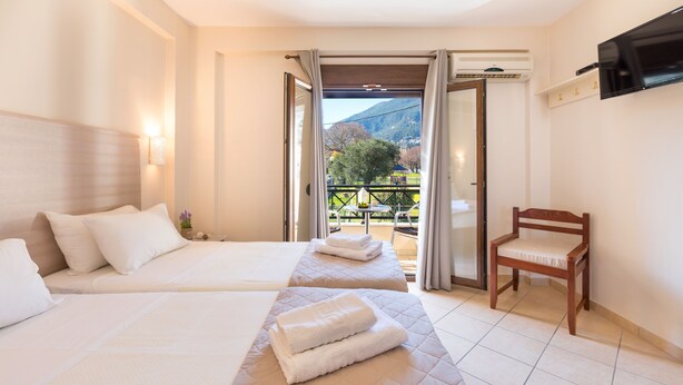 A hotel room with two beds, a chair, a TV, and a balcony with a mountain view. - Lemon Tree Corfu Apartments by Konnect (Corfu)