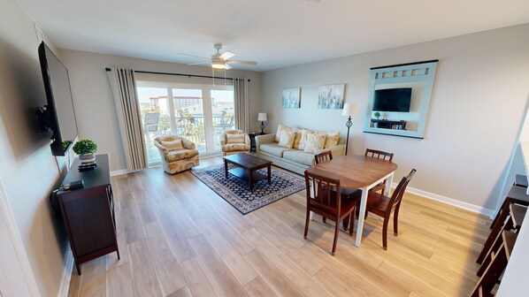 Un salon spacieux comprenant un canapé, des fauteuils, une table basse et une table à manger. Un téléviseur est fixé au mur, il y a un ventilateur de plafond et un balcon avec vue.