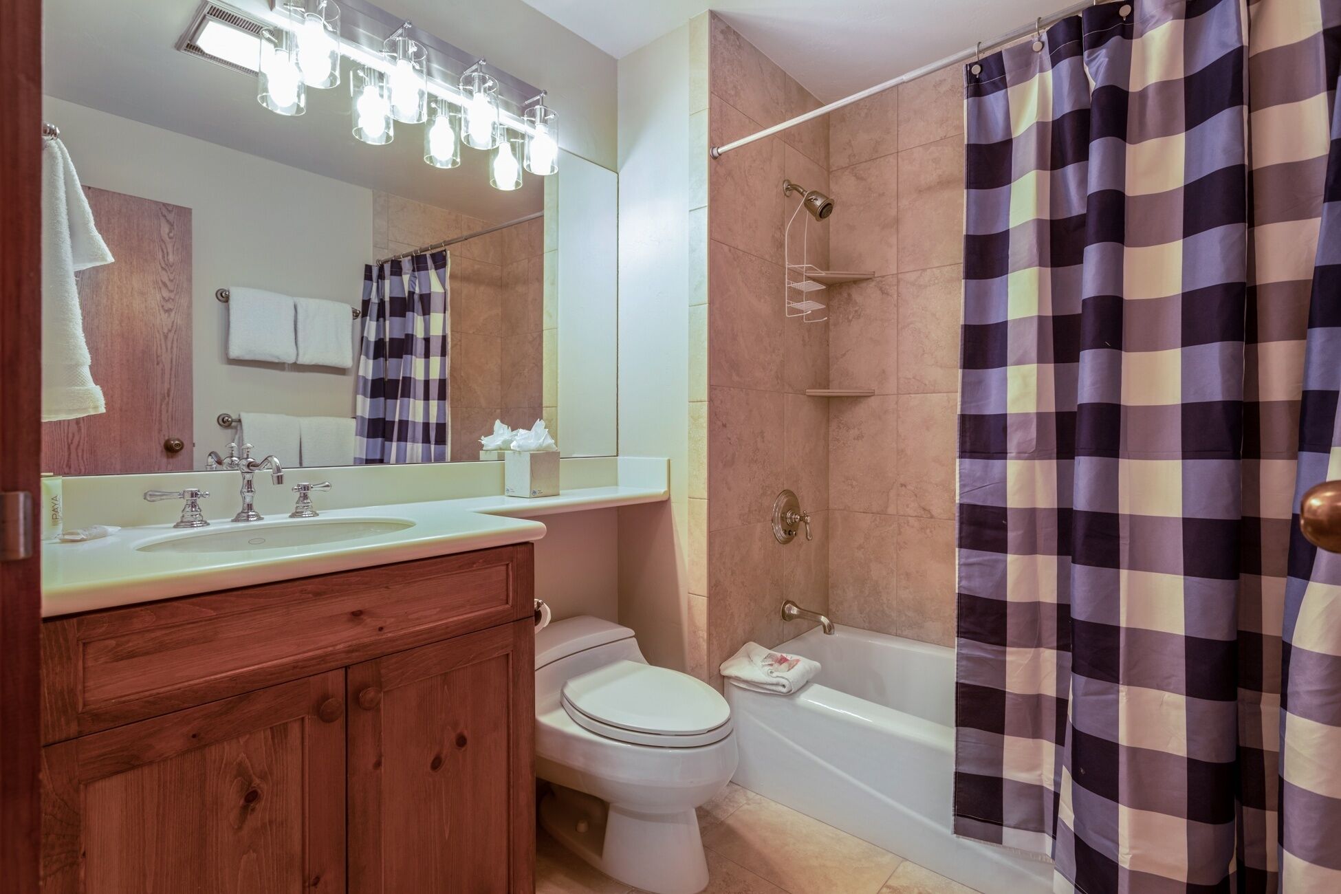 A bathroom with a wooden vanity, a sink, a toilet, a shower with a checkered curtain, and a mirror with lights above.