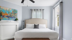 A bedroom with a large bed, a ceiling fan, a window with curtains, a painting on the wall, and a nightstand with a lamp. - The Blackberry Suite | 3 BR 2BA | 2nd Floor Condo (Charleston)