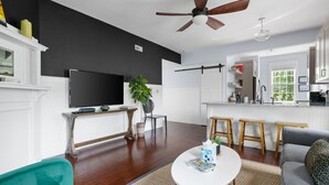A modern living room with a flat-screen TV, a ceiling fan, a dining area with stools, and a kitchen with white cabinets. - The Blackberry Suite | 3 BR 2BA | 2nd Floor Condo (Charleston)