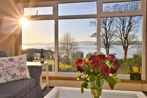 A room with a large window, a sofa, a table with a vase of roses, and a bottle of wine. - Tranquil location, waterside, pet friendly (Tighnabruaich, Otter Ferry)