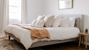 Un dormitorio con cama, almohadas, una mesita de noche y una ventana con cortinas.