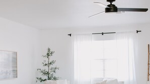 Una sala de estar moderna con ventilador de techo, muebles blancos, una mesa de centro de madera, un taburete redondo de madera y una planta decorativa.
