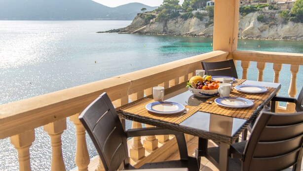 Une table dressée pour deux personnes, avec des assiettes, un saladier de fruits et une tasse, donnant sur une zone côtière où l’eau est claire et où l’on aperçoit une montagne au loin.