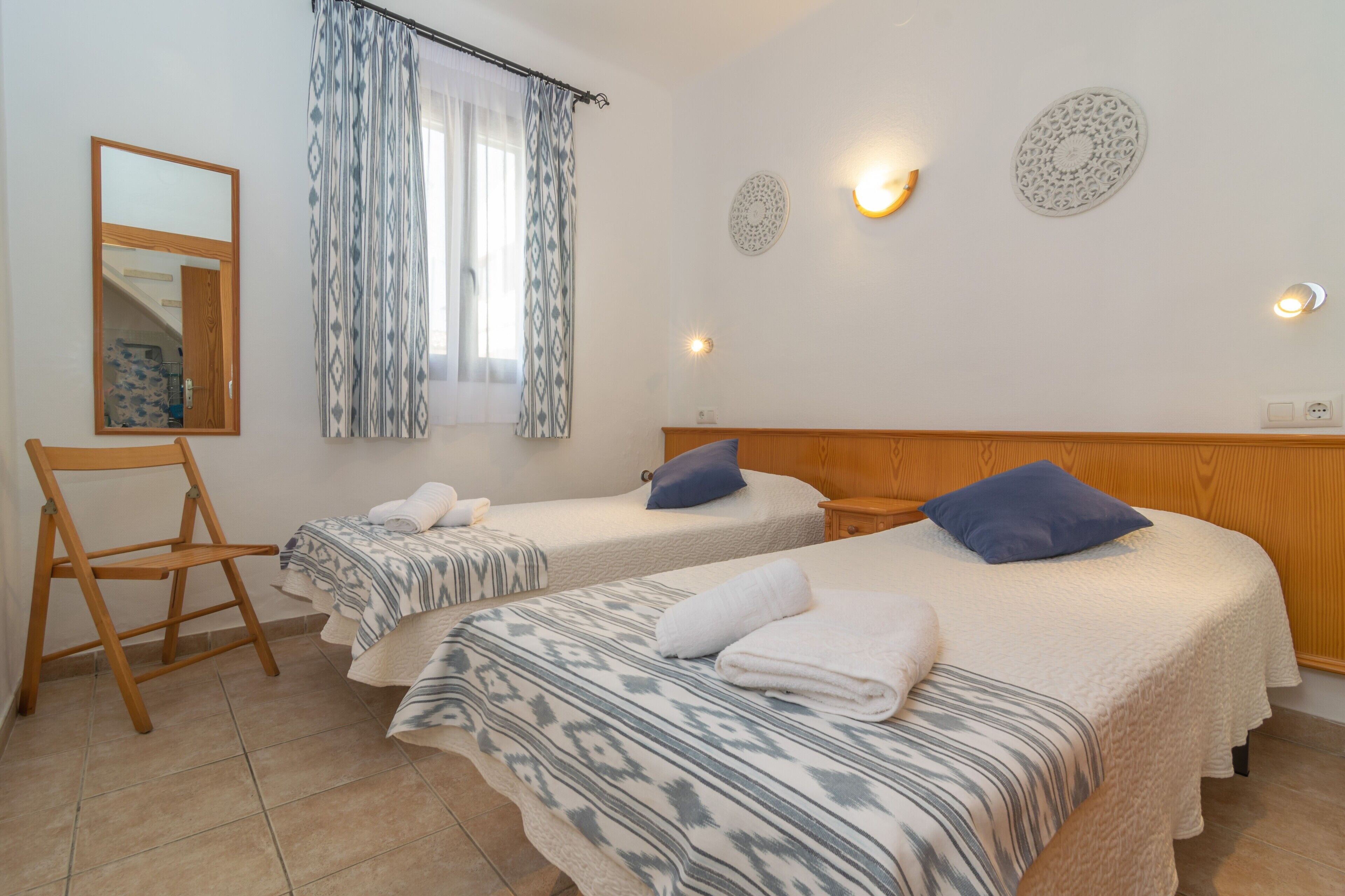 Una camera d'albergo con due letti, una sedia in legno, uno specchio, tende a motivi decorativi e luci a parete.
