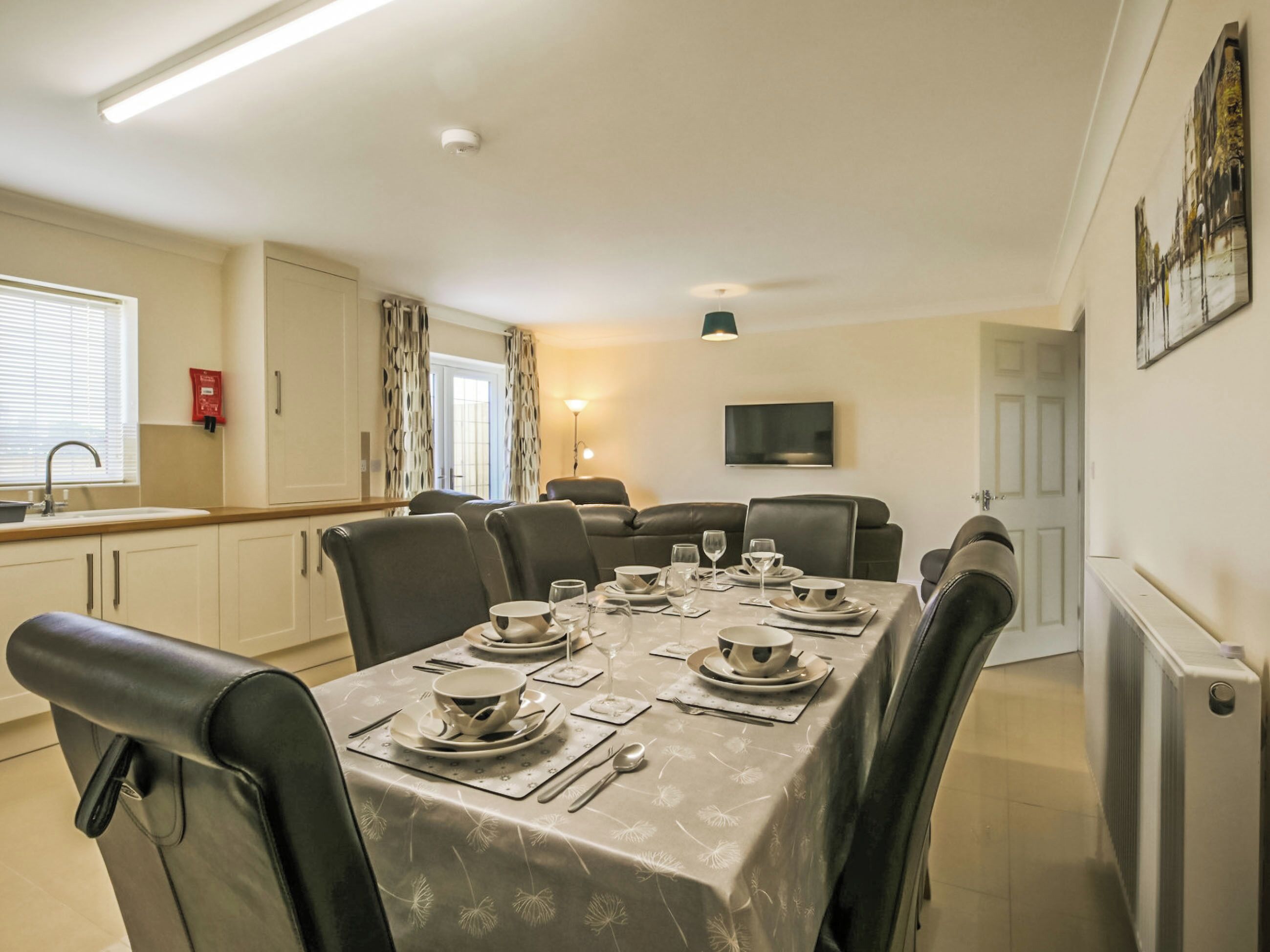 A dining area with a table set for six, a television mounted on the wall, and a window with curtains.