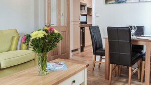 Ett modernt vardagsrum med en soffa, ett matbord med stolar, en vas med blommor och ett köksomr åde i bakgrunden.
