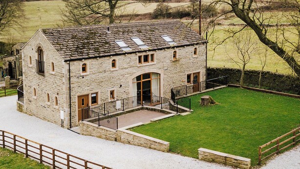 A stone building with a sloped roof, a central entrance, and a fenced courtyard. - Sycamore Barn - UK33353 (Northowram, near Halifax)