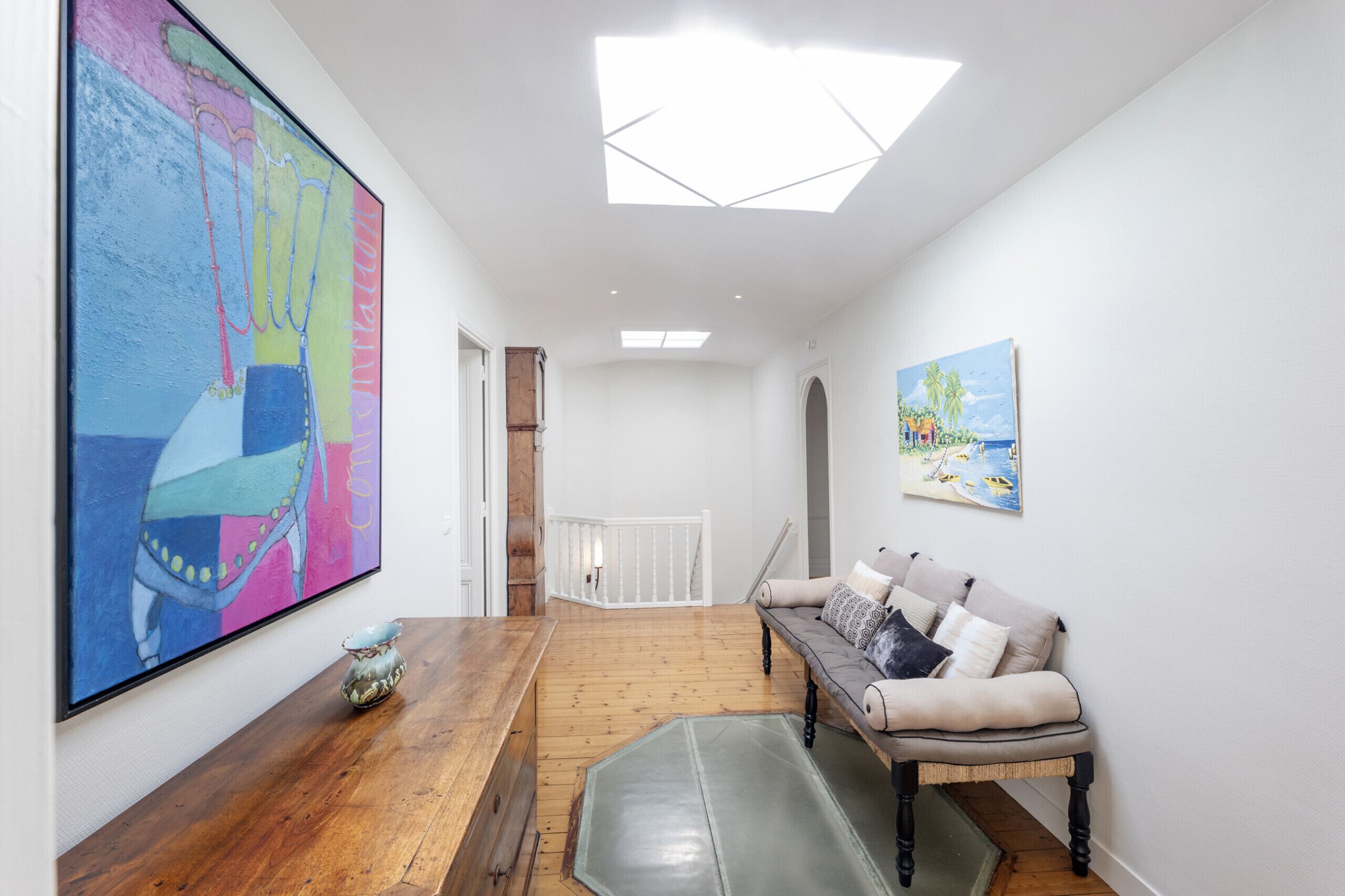 A hallway with a wooden floor, a bench, a painting, and a ceiling light.