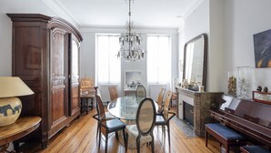 Un comedor con una mesa redonda de vidrio, sillas de madera, una araña y un piano.