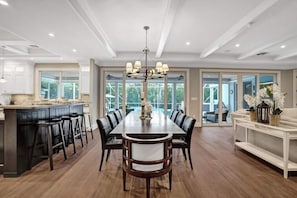 A spacious dining area with a long table and chairs, a kitchen area with a bar, and large windows offering views of the outdoors. - Luxe Modern Design 5000sf/Heated pool/Gameroom (Hollywood)
