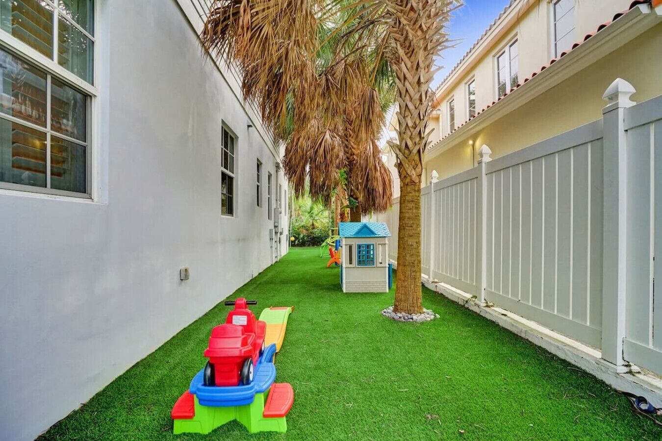 A narrow courtyard with artificial grass, a palm tree, a small playhouse, and a toy train.