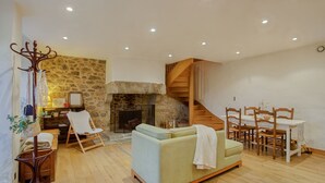 Ein gemütliches Wohnzimmer mit Kamin aus Stein, einer Holztreppe, einer Essecke mit Tisch und Stühlen sowie einem hellen Sofa.