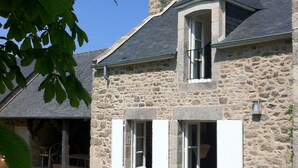 Ein Steinhaus mit geneigtem Dach, einer überdachten Terrasse mit Sitzgelegenheit und einer Steinsichtmauer, die das Grundstück umgibt.