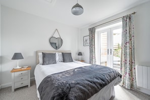 A bedroom with a bed, two bedside tables with lamps, a mirror, and a window with curtains. - Morley's (Truro)