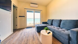 Een moderne woonkamer met een grijze bank, een ronde witte salontafel en uitzicht op het strand door een glazen deur.