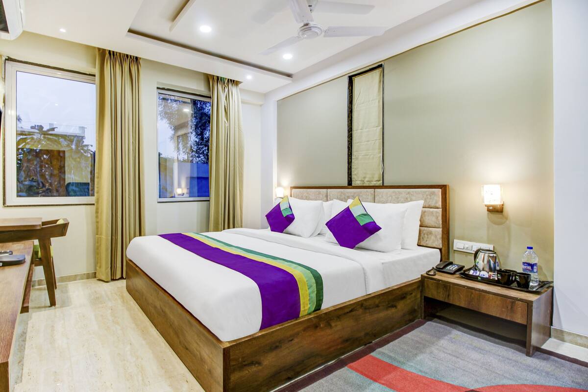Ein Hotelzimmer mit einem hölzernen Bett, einem Schreibtisch, einem Stuhl, einem Fernseher, einem Fenster mit Vorhängen und einem Nachttisch mit Lampe und Flasche.