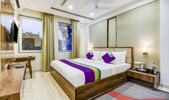 Habitación de hotel con una cama de madera, un escritorio, una silla, un televisor, una ventana con cortinas y una mesita de noche con una lámpara y una botella.