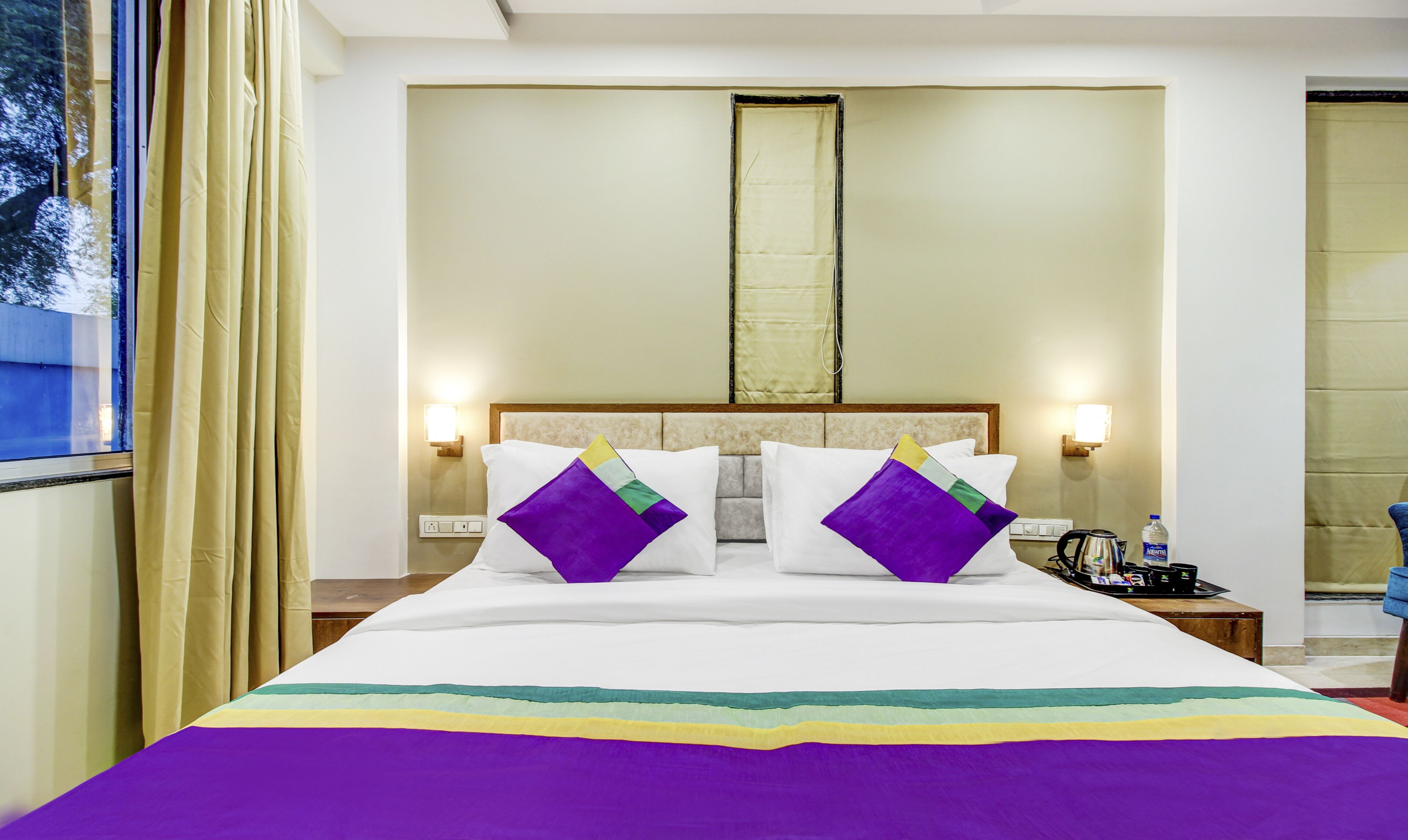 A hotel room with a large bed, two bedside tables, a nightstand, a window with curtains, and a view of trees outside.