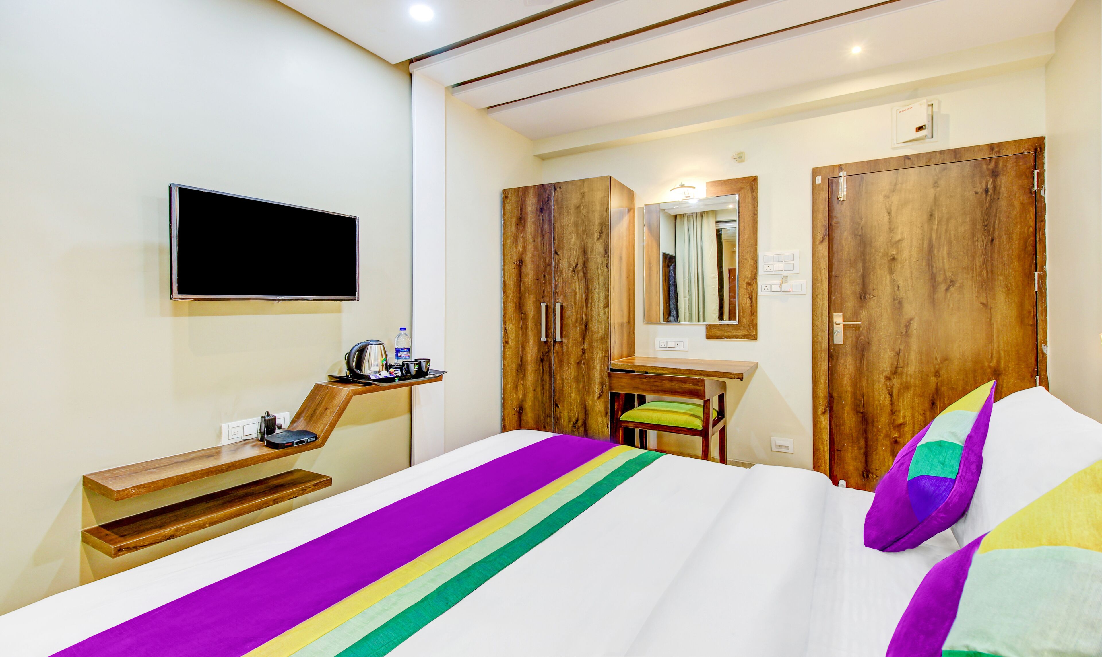 Habitación de hotel con cama, armario de madera, escritorio, espejo y televisor montado en la pared.