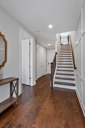 Un pasillo con suelo de madera, paredes blancas y una escalera de madera con barandilla.