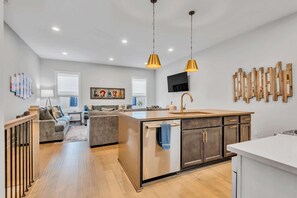 A modern kitchen with a central island, pendant lighting, and a dining area with a sofa and a TV.