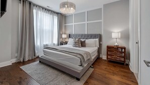 A bedroom with a large bed, a chandelier, a nightstand, and a view of the outside. - Two Luxe Nashville Homes + Downtown Skyline Views (Nashville)