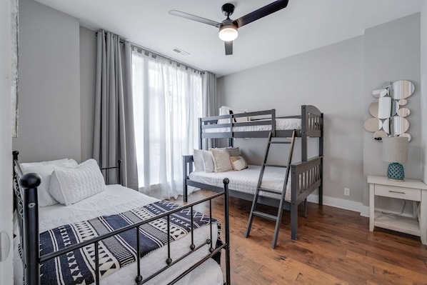 A bedroom with a bunk bed, a single bed, a ceiling fan, a bedside table, and a wall-mounted mirror.
