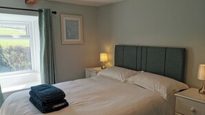 Ein ordentlich bezogenes Bett mit Kissen und gefalteten Decken, eine Nachttischlampe, ein gerahmtes Bild an der Wand und ein Fenster mit Vorhängen.