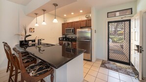 A modern kitchen with a central island, wooden cabinets, stainless steel appliances, pendant lighting, and a dining area with wooden chairs. - Grand Champions Villas 61- Wailea (Kihei)