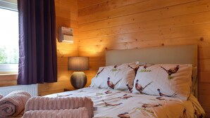 A wooden cabin room with a bed, pillows with bird prints, a lamp, and a radiator. - Pheasant Lodge - UK33402 (Billesdon, near Leicester)