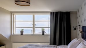 Un dormitorio con un ventanal grande, una cama con ropa blanca, un edredón gris y dos macetas con plantas en el alféizar.