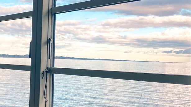 Vista a través de una ventana con marco metálico, que da a una masa de agua y una costa distante con nubes en el cielo.