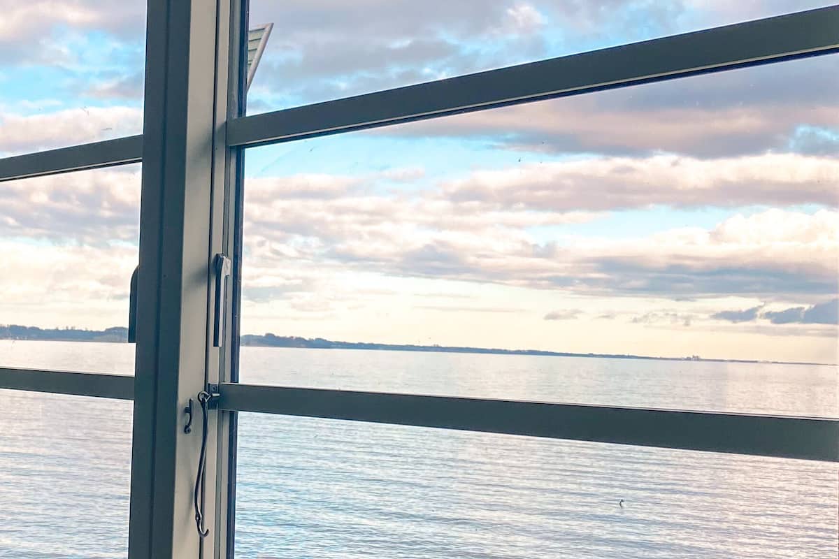 Vista a través de una ventana con marco metálico, que da a una masa de agua y una costa distante con nubes en el cielo.
