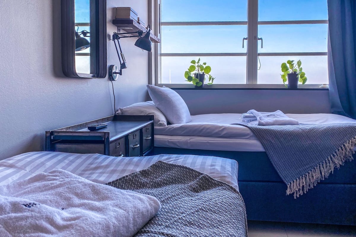 Una habitación de hotel con cama, mesita de noche, lámpara y ventana con plantas.